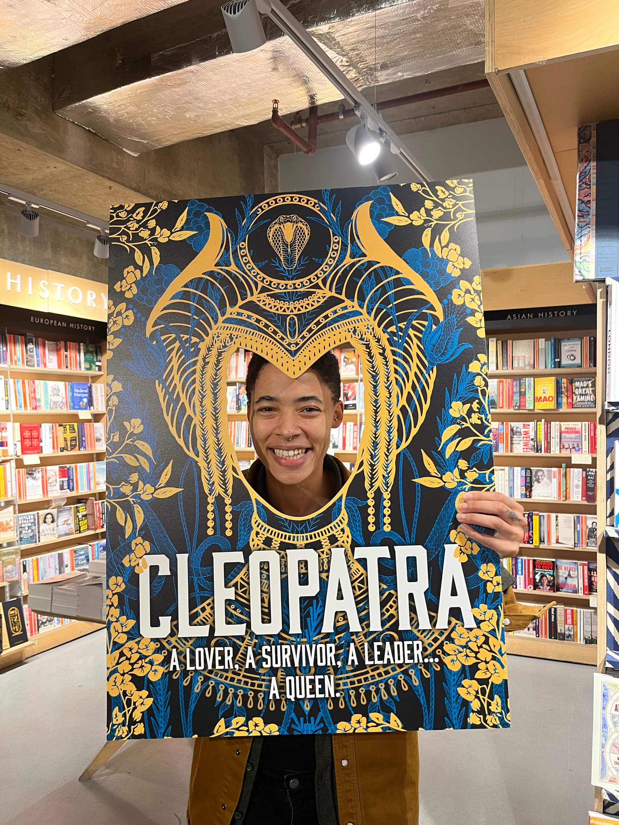C. L. Clark with their face in a cut-out frame version of Cleopatra the book cover, with their face where the face of Cleopatra would be.