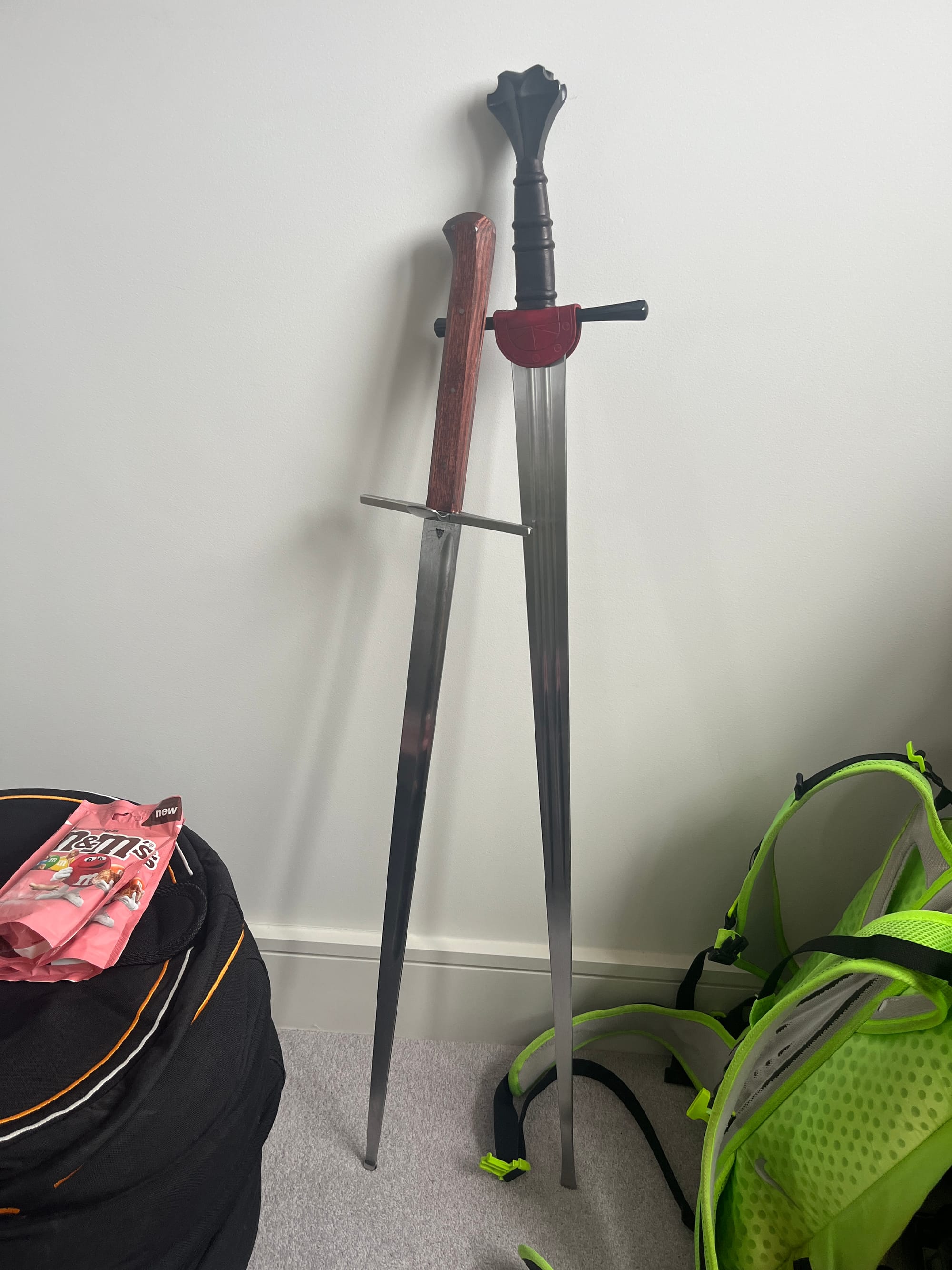 Two swords leaning up against each other against a white wall. Ill advised. Cookie dough M&Ms on a sandbag next to a running backpack.